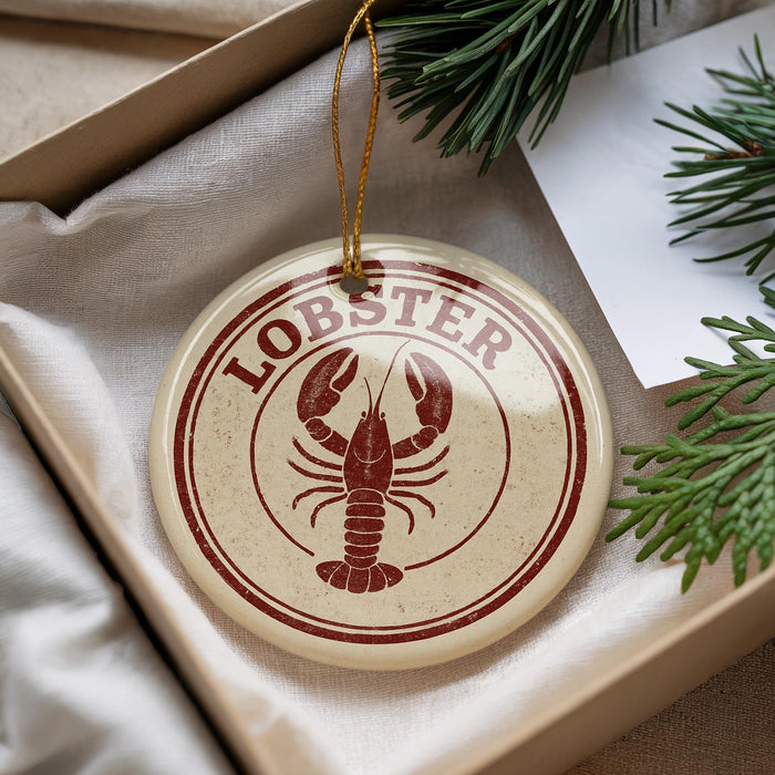 A round ornament with a lobster design is placed inside a box, surrounded by pine branches.