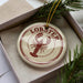 A round ornament with the word "LOBSTER" printed on it, featuring a lobster design, is displayed in a box with pine branches.