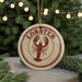 A round ornament with a lobster design, featuring the word "LOBSTER" in red text, is hanging from a string in front of a Christmas tree with twinkling lights.