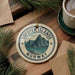 A circular ornament with the text "Lofoten Islands NORWAY" and a mountain landscape design, hanging on a wooden surface.