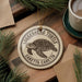 A circular ornament featuring a turtle design, with the text "Loggerhead Turtle" and "Caretta Caretta" printed on it, is displayed on a wooden surface alongside a cup of coffee and pine branches.