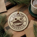 A circular ornament with the text "LOMBOK INDONESIA" printed on it, surrounded by pine branches and a cup of coffee on a wooden surface.