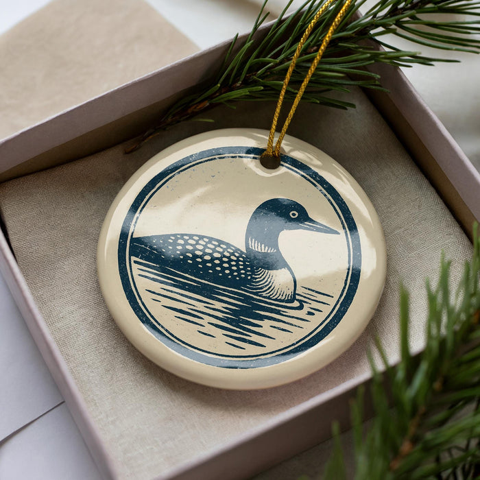 A ceramic ornament in the shape of a loon, featuring a blue and white design, is displayed in a box with pine branches.