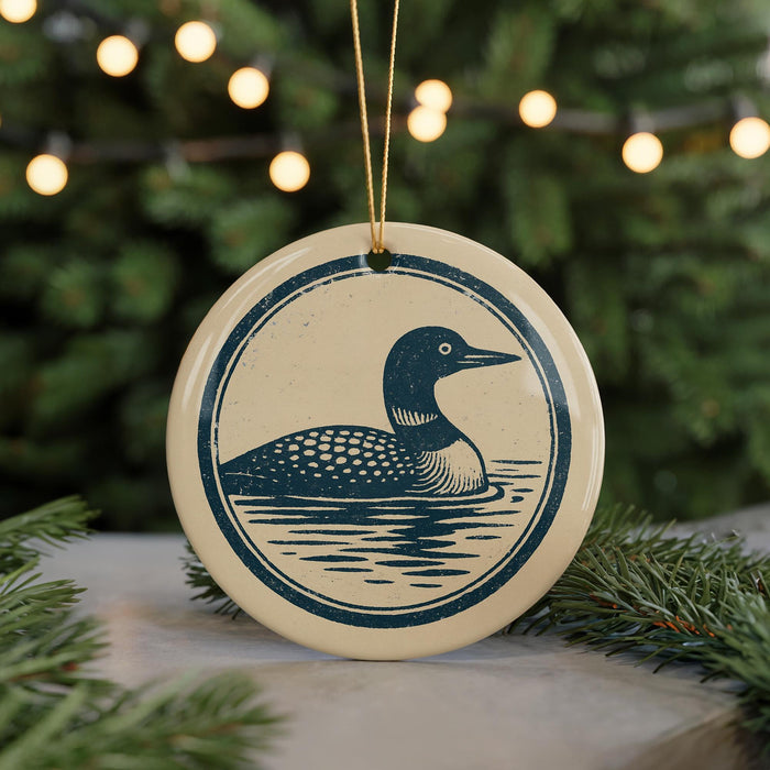 A round ceramic ornament featuring a loon, a type of bird, is displayed in front of a blurred background with Christmas lights.