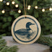 A round ceramic ornament featuring a loon, a type of bird, is displayed in front of a blurred background with Christmas lights.