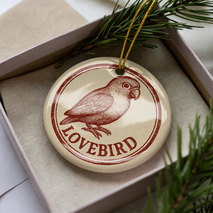 A round ornament with a bird image and the word "LOVEBIRD" printed on it, placed inside a box with pine branches.