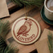 A round ornament with a bird image and the word "LOVEBIRD" printed on it, placed on a wooden surface next to a cup of coffee and pine branches.