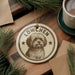 A circular ornament featuring a dog's portrait, with the text "LÖWCHEN" prominently displayed. The ornament is hanging from a string, and there is a cup of coffee and some pine branches in the background.