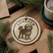 A circular ornament featuring a dog illustration, with the word "LÜSChen" written around the edge. The ornament is placed on a wooden surface, with pine branches and a cup of coffee nearby.