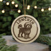 A circular ornament featuring a dog illustration and the text "LÖWCHEN" (Lowchen) on it, hanging in front of a Christmas tree with twinkling lights.