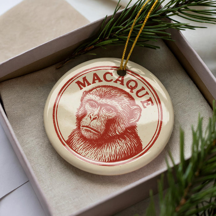 A round ornament with a monkey's face on it, placed inside a box with pine branches.