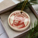 A round ornament with a monkey's face on it, placed inside a box with pine branches.