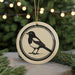 A round ceramic ornament featuring a black and white illustration of a bird, likely a magpie, is displayed in front of a blurred background of Christmas lights.