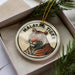 A circular ornament featuring a tiger's image, placed in a box with pine branches.