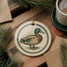 A ceramic ornament featuring a duck is displayed on a wooden surface, accompanied by a cup of coffee and some pine branches.