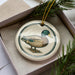 A ceramic ornament in the shape of a duck is displayed in a box, surrounded by pine branches.