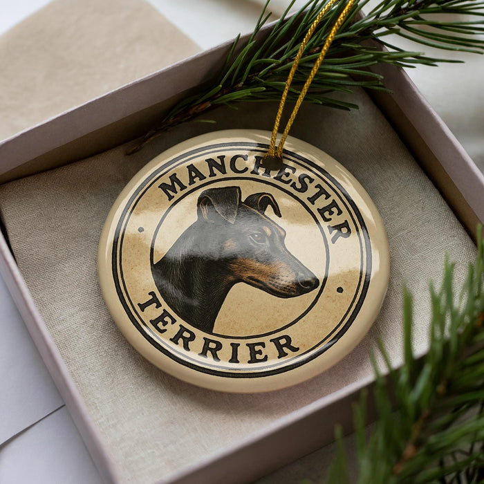 A circular ornament with a dog's head and the text "MANCHESTER TERRIER" printed on it, placed inside a box with pine branches.