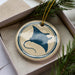 A round ceramic ornament with a blue and white design depicting a stingray or manta ray, hanging from a string.