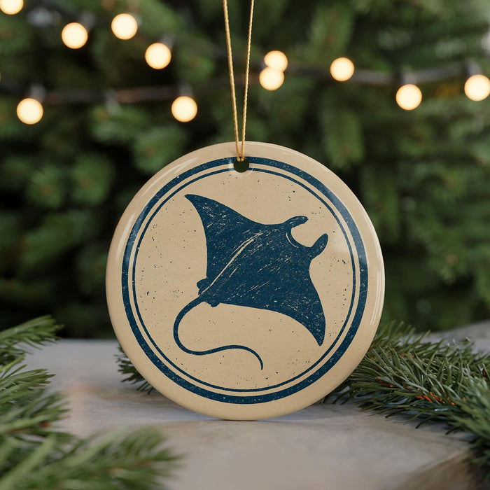 A round ornament with a blue and white design featuring a stingray silhouette is displayed against a blurred background of a Christmas tree with twinkling lights.
