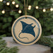 A round ornament with a blue and white design featuring a stingray silhouette is displayed against a blurred background of a Christmas tree with twinkling lights.