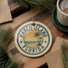 A circular ornament with the text "MARCO ISLAND FLORIDA" and a palm tree design, placed on a wooden surface next to a cup of coffee and some pine branches.