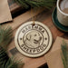 A circular ornament with the text "Maremma Sheepdog" and a graphic of a dog's head is displayed on a wooden surface, surrounded by pine branches and a cup of coffee.
