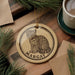 A round ornament featuring a leopard cat, with the name "Margay" written on it, is displayed on a wooden surface alongside a cup of coffee and some pine branches.