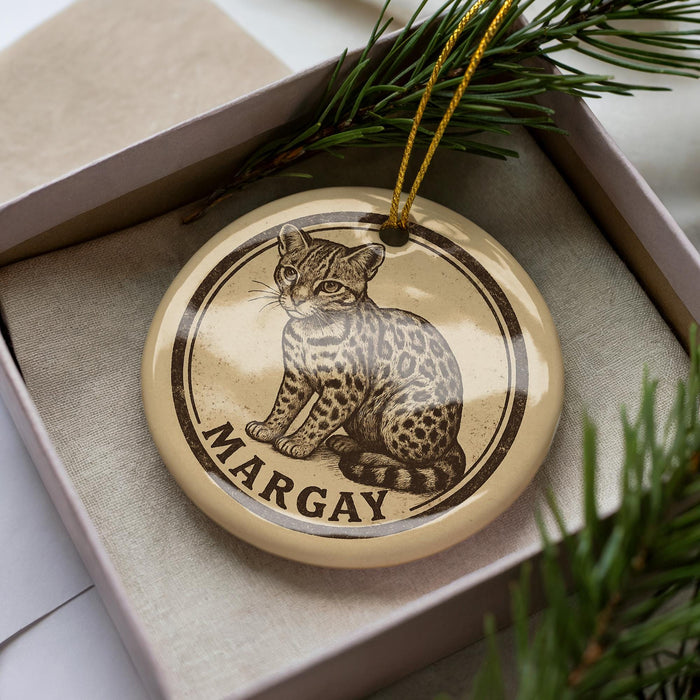 A round ornament with a leopard print design and the name "Margay" printed on it, placed inside a box with pine branches.