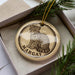A round ornament with a leopard print design and the name "Margay" printed on it, placed inside a box with pine branches.