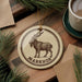 A circular ornament featuring a goat with horns, placed on a wooden surface next to a cup of coffee and pine branches.