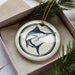 A white ceramic ornament with a blue and black design depicting a fish is displayed in a box, surrounded by pine branches.
