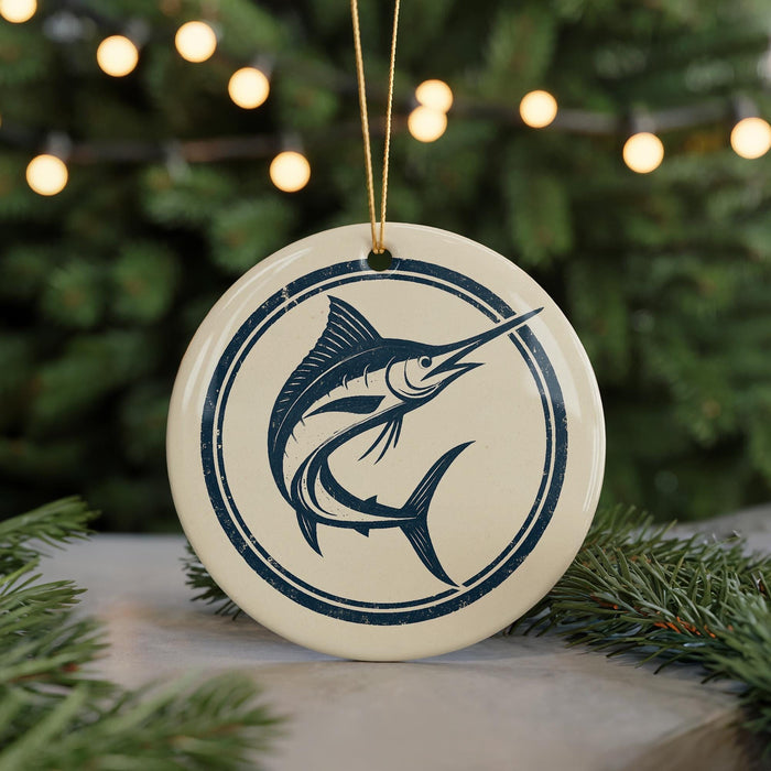 A round ornament featuring a blue and white illustration of a swordfish is displayed against a blurred background of Christmas lights and pine branches.
