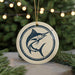 A round ornament featuring a blue and white illustration of a swordfish is displayed against a blurred background of Christmas lights and pine branches.