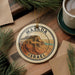 A circular ornament with the text "MASADA ISRAEL" and an illustration of a mountain range, placed on a wooden surface next to a cup of coffee and pine branches.