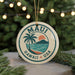 A circular ornament with the text "MAUI HAWAII USA" and a palm tree design, hanging from a string against a background of Christmas lights and greenery.