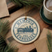 A circular ornament with the text "Memphis, Tennessee, USA" printed on it, surrounded by pine branches and a cup of coffee on a wooden surface.