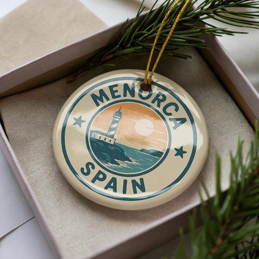 A circular ornament with the text "MENORCA SPAIN" and a lighthouse image, placed in a box with pine branches.