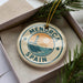 A circular ornament with the text "MENORCA SPAIN" and a lighthouse image, placed in a box with pine branches.