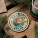 A circular ornament with the text "MENORCA SPAIN" and an illustration of a lighthouse is displayed on a wooden surface, surrounded by pine branches and a cup of coffee.