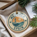 A circular ornament with the text "MENORCA SPAIN" and an illustration of a lighthouse on a beach, hanging from a string.