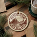 A circular ornament with the text "Milford Sound, New Zealand" printed on it, surrounded by pine branches and a cup of coffee on a wooden surface.