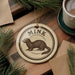A round ornament with the word "MINK" printed on it, featuring a drawing of a brown animal, is displayed on a wooden surface alongside a cup of coffee and some pine branches.