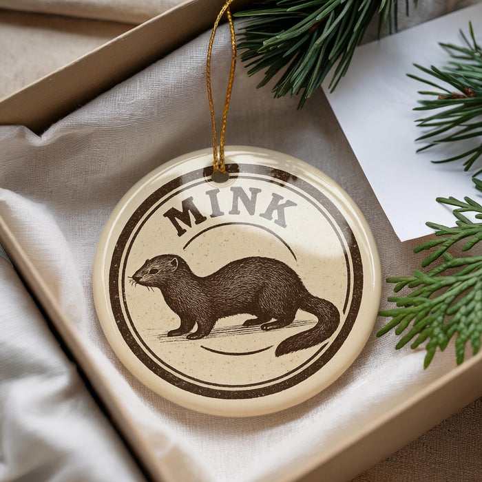 A round ornament with a mink illustration on it, placed in a box with pine branches.