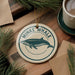 a round ornament with a blue and white design depicting a whale, hanging on a wooden surface. There is also a cup of coffee and some pine branches in the background.