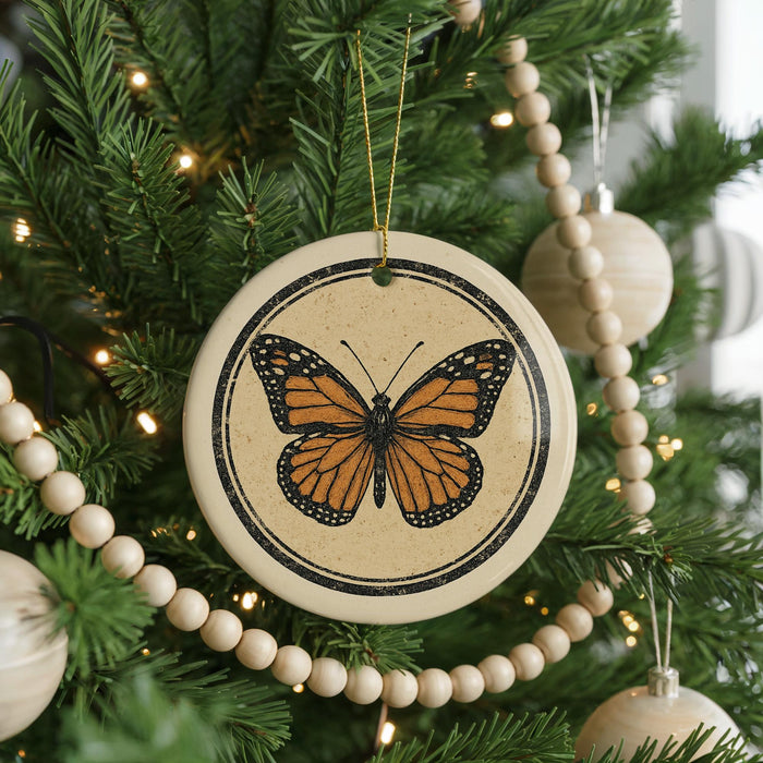 A ceramic ornament featuring a detailed image of a butterfly is hanging from a Christmas tree, surrounded by other ornaments and lights.