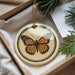A decorative ornament featuring a butterfly is displayed in a box, surrounded by pine branches and greenery.