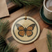 a round ceramic ornament with a butterfly design, placed on a wooden surface alongside a cup of coffee and some pine branches.