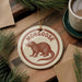 A circular ornament featuring a brown mongoose is displayed on a wooden surface, surrounded by pine branches and a cup of coffee.