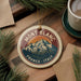 A round ornament with a mountain and text, likely representing the location of Mont Blanc, France, is displayed on a wooden surface alongside a cup of coffee and some pine branches.