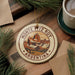 A round ornament with a mountain landscape and text, hanging on a wooden surface next to a cup of coffee and some pine branches.